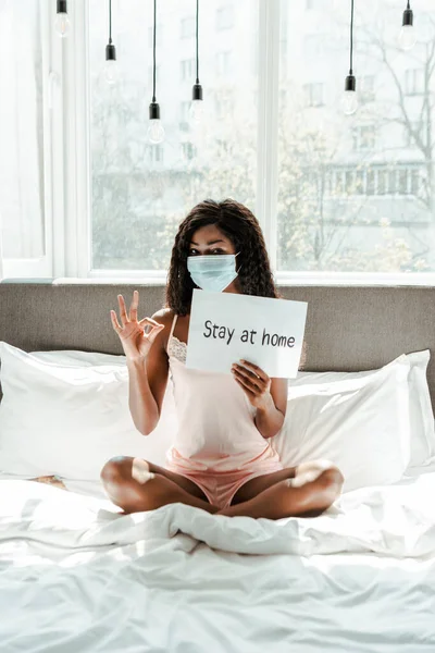 Africano americano mulher em máscara médica com pernas cruzadas mostrando bem sinal e cartaz com estadia em casa inscrição na cama no quarto — Fotografia de Stock