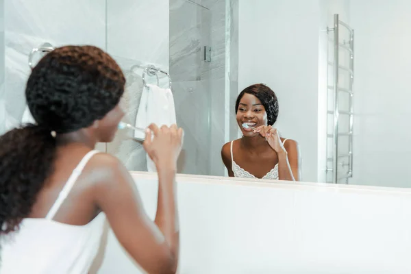 Mujer afroamericana sonriendo, cepillándose los dientes y mirando el espejo en el baño - foto de stock