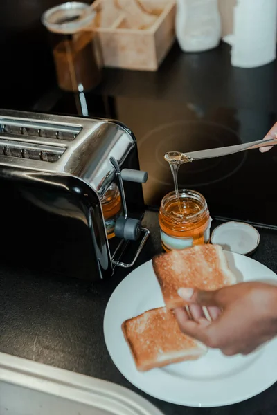 Ausgeschnittene Ansicht einer afrikanisch-amerikanischen Frau bei der Zubereitung von Toast mit Honig in der Nähe von Toaster in der Küche — Stockfoto