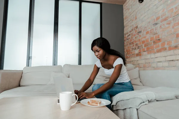 African american freelancer using laptop at coffee table with cup of tea and plate with toasts in living room — Stock Photo