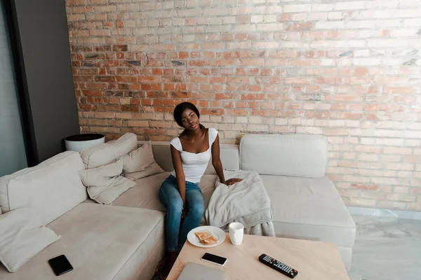 Hochwinkelblick einer afrikanisch-amerikanischen Frau auf einem Sofa in der Nähe eines Couchtisches mit Teller, Tasse Tee, Smartphone und Fernbedienung im Wohnzimmer — Stockfoto