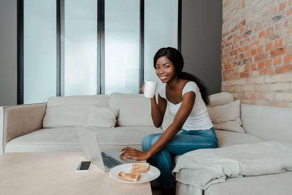 Freelancer afroamericano con taza de té sonriendo y usando portátil en la mesa de café con smartphone y plato con tostadas en la sala de estar - foto de stock