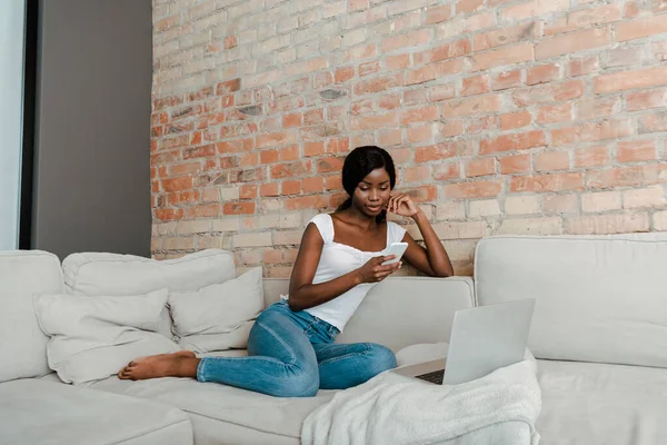 African american freelancer using smartphone near laptop on sofa in living room — Stock Photo