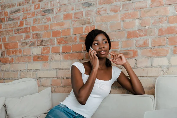 Africano menina americana falando no smartphone perto da parede de tijolo no sofá na sala de estar — Fotografia de Stock