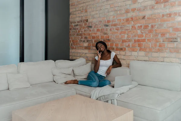 Freelancer afroamericano hablando en smartphone y sonriendo en sofá en la sala de estar - foto de stock