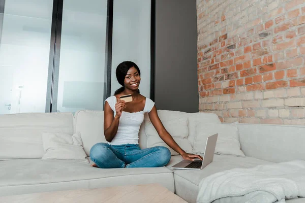 Afroamerikanerin lächelt, schaut in die Kamera, zeigt Kreditkarte und benutzt Laptop auf Sofa im Wohnzimmer — Stockfoto