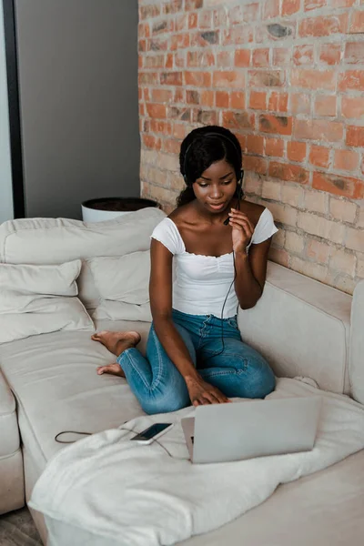 Vista de alto ângulo do freelancer afro-americano no fone de ouvido perto do laptop e smartphone no sofá na sala de estar — Fotografia de Stock