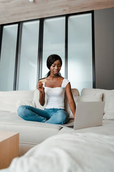African american woman looking at laptop and showing credit card on sofa in living room — Stock Photo