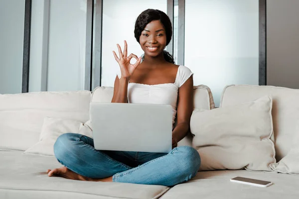 Freelancer afroamericano con piernas cruzadas y portátil sonriendo y mostrando el cartel de bien cerca del teléfono inteligente en el sofá en la sala de estar - foto de stock