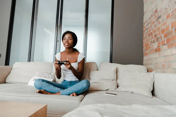 KYIV, UKRAINE 10 AVRIL 2020 : femme afro-américaine aux jambes croisées jouant au jeu vidéo avec joystick sur canapé dans le salon — Photo de stock