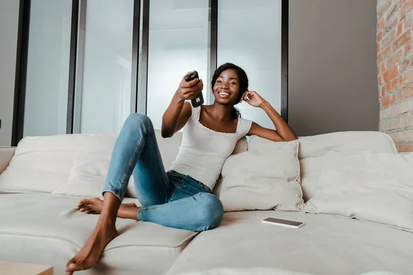 Mujer afroamericana sonriendo con controlador remoto cerca de teléfono inteligente en el sofá en la sala de estar - foto de stock