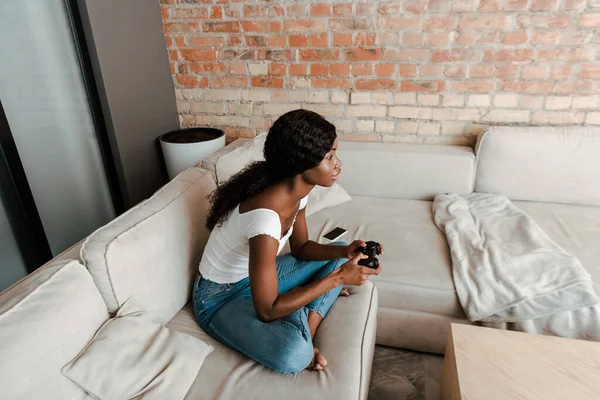 KYIV, UCRAINA 10 APRILE 2020: vista ad alto angolo della donna afroamericana con le gambe incrociate che gioca al videogioco con joystick sul divano in salotto — Foto stock