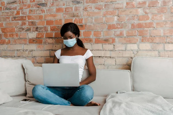 African american freelancer in medical mask with crossed legs working on laptop near smartphone on sofa in living room — Stock Photo