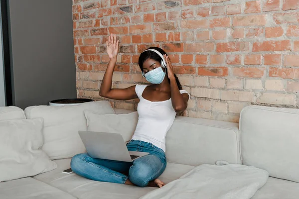 African american freelancer in medical mask with hand in air, headphones and laptop on sofa in living room — Stock Photo