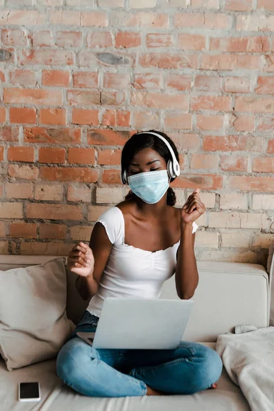 Африканский фрилансер в медицинской маске и наушниках слушает музыку с ноутбука возле смартфона на диване в гостиной — стоковое фото