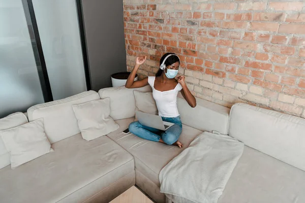 Vue grand angle de pigiste afro-américain en masque médical et écouteurs écoutant de la musique avec ordinateur portable sur canapé dans le salon — Photo de stock