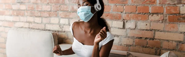 Coltivazione orizzontale di donna afro-americana con maschera medica ascoltare musica in cuffie vicino muro di mattoni in soggiorno — Foto stock