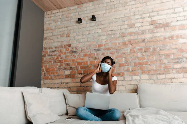 Африканский фрилансер в медицинской маске и наушниках слушает музыку с ноутбука рядом со смартфоном на диване — стоковое фото