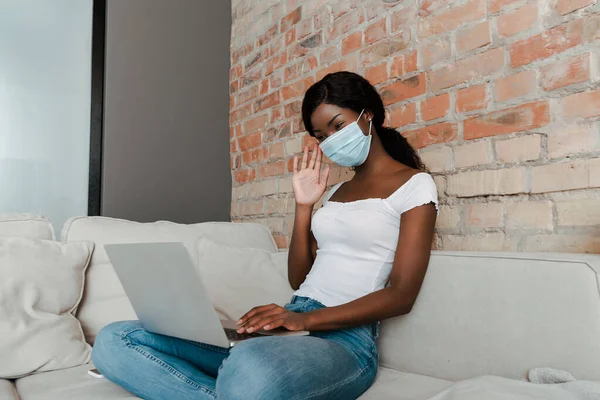 Libero professionista afroamericano in maschera medica con videochiamata sul divano in soggiorno — Foto stock