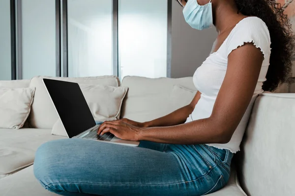 Vista recortada del freelancer afroamericano en máscara médica trabajando con portátil en sofá en sala de estar - foto de stock