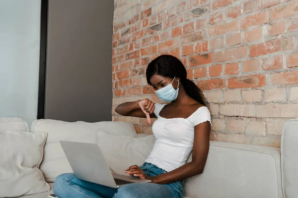 Freelancer afroamericano en máscara médica con piernas cruzadas y portátil mostrando signo de aversión en el sofá en la sala de estar - foto de stock