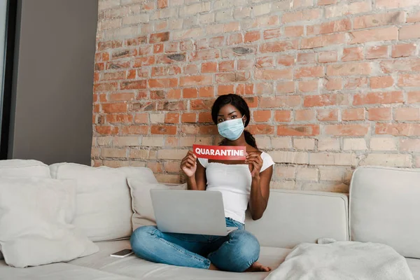 Afroamerikanischer Freiberufler in medizinischer Maske mit überkreuzten Beinen und Laptop zeigt Karte mit Quarantäne-Schriftzug auf Sofa im Wohnzimmer — Stockfoto