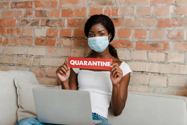 African american freelancer in medical mask with laptop showing card with quarantine lettering and looking at camera on sofa — Stock Photo