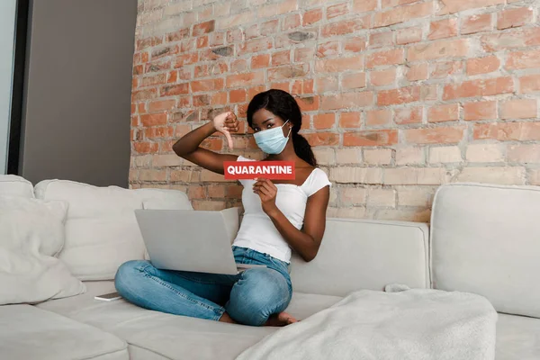 Freelancer afroamericano en máscara médica con portátil mostrando tarjeta con letras de cuarentena y signo de aversión en el sofá - foto de stock