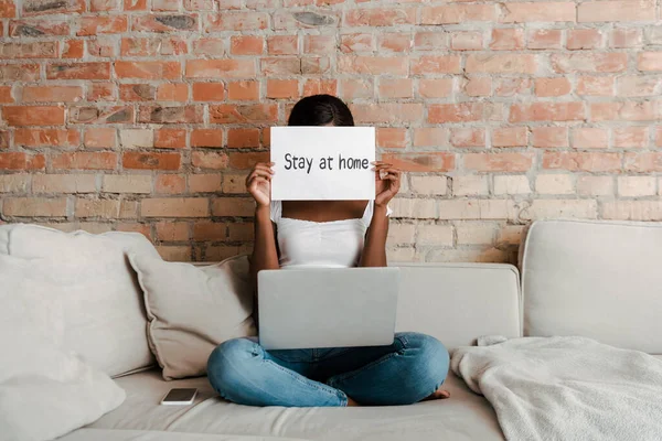 African american freelancer with crossed legs and laptop showing placard with stay at home lettering on sofa in living room — Stock Photo