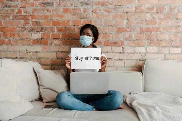 African american freelancer in medical mask with laptop near smartphone showing placard with stay at home lettering on sofa in living room — Stock Photo