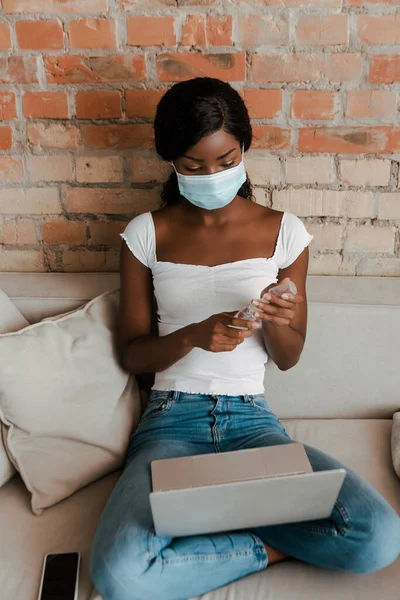 African american freelancer in medical mask with laptop near smartphone holding hand sanitizer on sofa in living room — Stock Photo