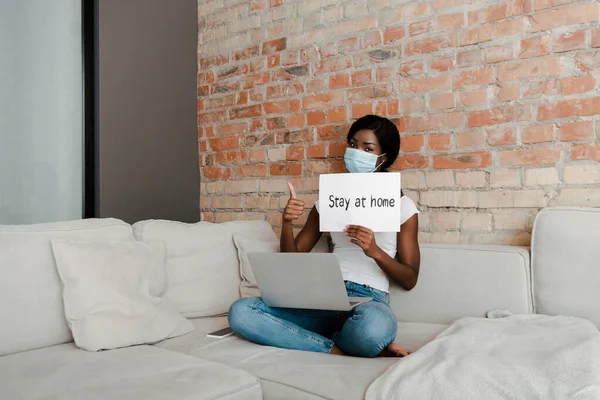 Freelancer afroamericano en máscara médica con portátil mostrando como signo y cartel con la estancia en casa letras en el sofá - foto de stock