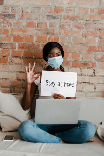 Pigiste afro-américain dans un masque médical avec ordinateur portable montrant signe correct et pancarte avec rester à la maison lettrage sur canapé dans le salon — Photo de stock