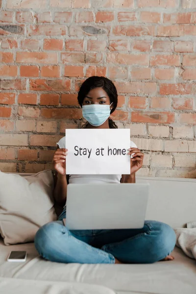 Freelancer afroamericano en máscara médica con portátil mostrando pancarta con letras en casa cerca de teléfono inteligente en sofá en la sala de estar - foto de stock