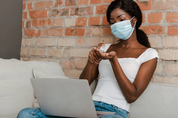Afro-américain pigiste dans un masque médical avec ordinateur portable en utilisant un désinfectant pour les mains sur le canapé dans le salon — Photo de stock