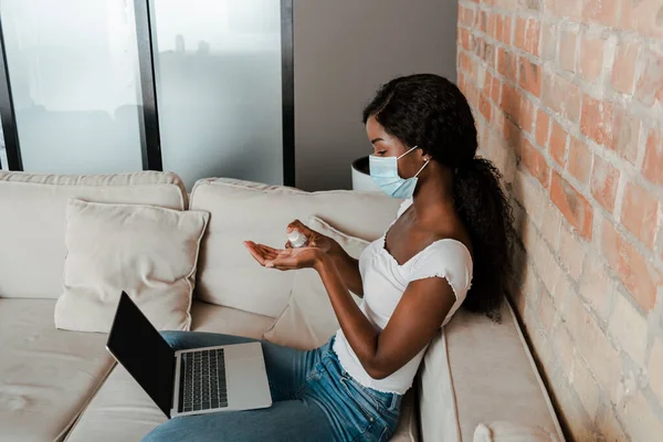 Високий кут зору афроамериканського фрілансера в медичній масці з ноутбуком, використовуючи ручний дезінфікуючий засіб на дивані у вітальні — стокове фото