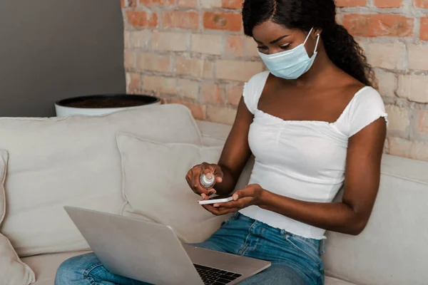 Libero professionista afroamericano in maschera medica con smartphone disinfettante portatile con disinfettante per le mani sul divano in soggiorno — Foto stock