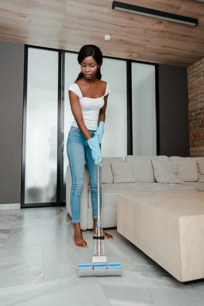 African american woman in rubber gloves cleaning up with mop in living room — Stock Photo