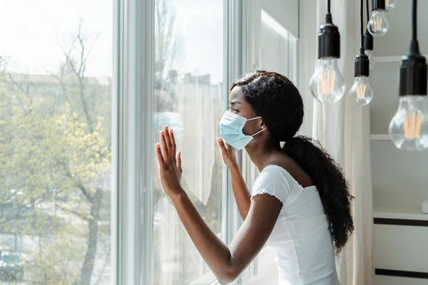 Africano americano mulher em máscara médica tocando janelas na sala de estar — Fotografia de Stock