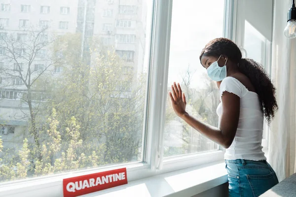 Chica afroamericana en máscara médica tocando ventanas cerca de la tarjeta con letras de cuarentena en la sala de estar - foto de stock