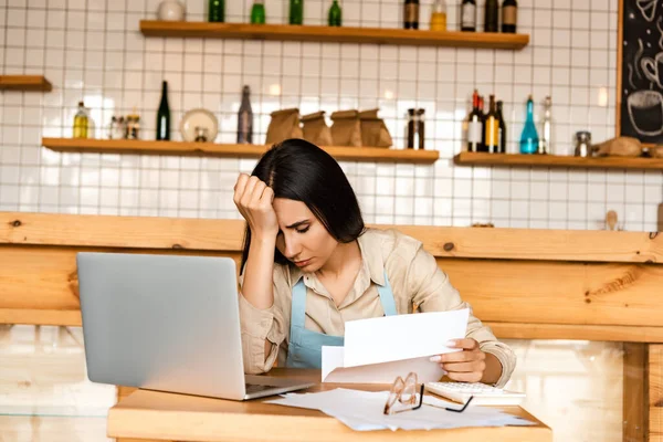 Proprietario del caffè sconvolto che tiene carta vicino alla calcolatrice, laptop e documenti con occhiali a tavola — Foto stock