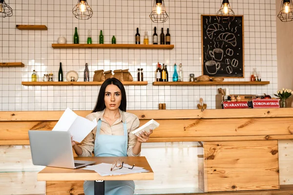 Cafe owner looking at camera and holding paper with calculator near laptop and documents with glasses at table — Stock Photo