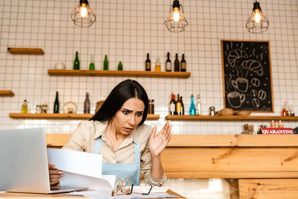 Shocked propietario de la cafetería con papeles cerca de la computadora portátil y gafas mirando la calculadora en la mesa - foto de stock