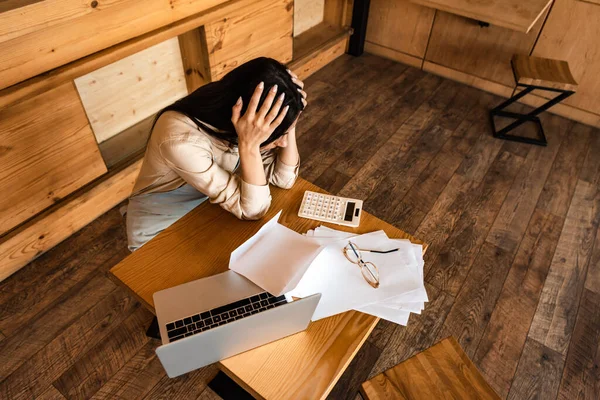 Vue aérienne du propriétaire du café touchant la tête près de l'ordinateur portable, des documents, des lunettes et de la calculatrice à table — Photo de stock