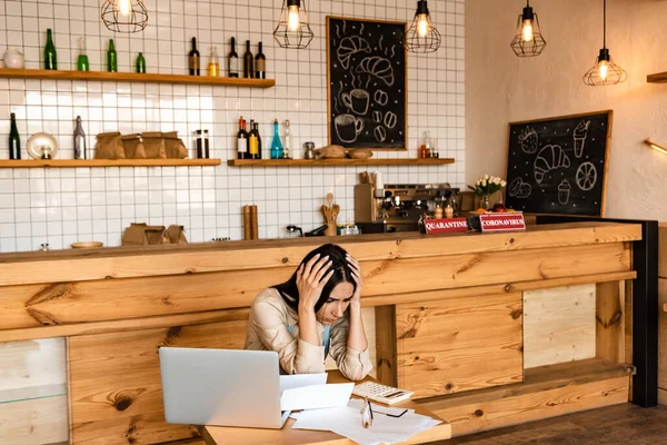 Propietario molesto café tocando la cabeza cerca de la computadora portátil, documentos y calculadora en la mesa - foto de stock