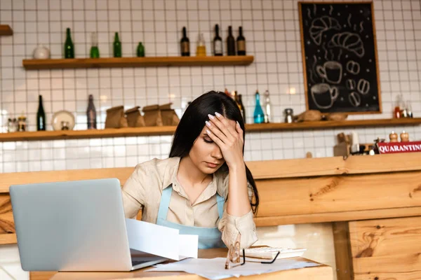Upset cafe owner covering face near laptop, documents and glasses at table — Stock Photo