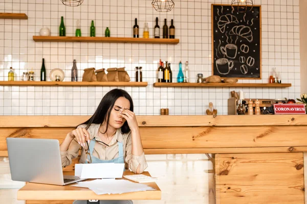 Cansado proprietário do café segurando óculos perto de calculadora, papéis e laptop na mesa — Fotografia de Stock