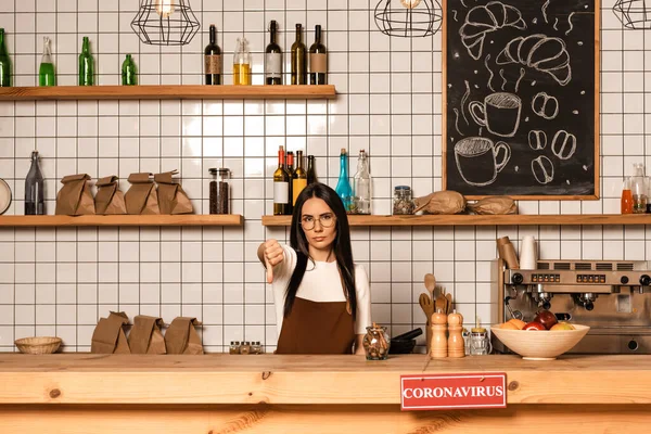 Brunette cafe owner looking at camera and showing dislike sign near table with card with coronavirus inscription — Stock Photo