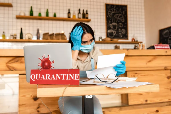 Proprietario di caffè preoccupato in documento di lettura maschera medica vicino a carte, laptop e carta con lettering quarantena a tavola — Foto stock