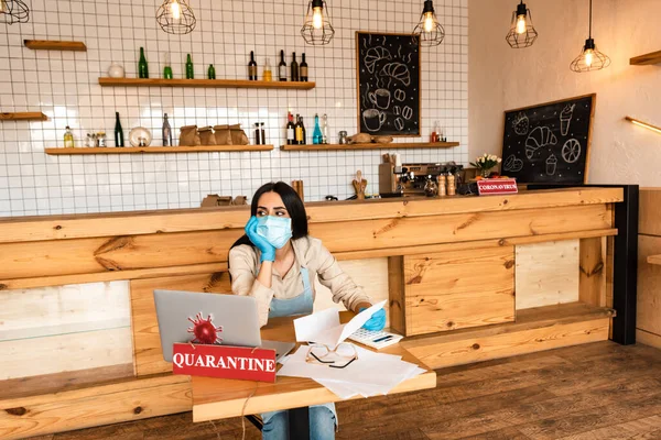 Propietario del café con máscara médica mirando hacia otro lado y sosteniendo papel cerca de la calculadora, documentos, computadora portátil y tarjeta con letras de cuarentena en la mesa - foto de stock